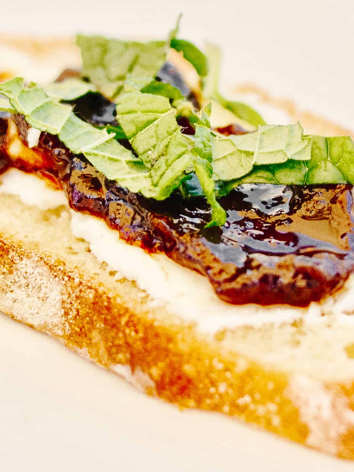 Close up of mint leave slices on top of fig jam, goat cheese and toasted baguette slices on white surface.