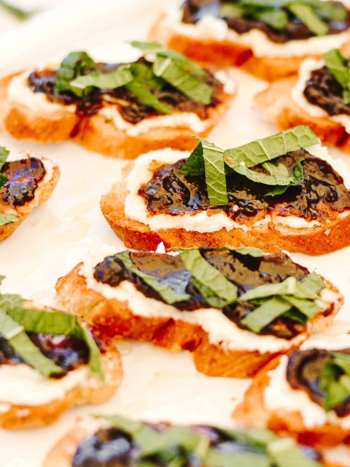 Plate full of toasted baguette slices, topped with cheese, jam and mint leaves.