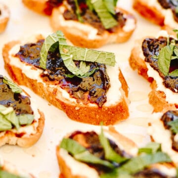 Plate full of slices of baguette with soft goat cheese spread on top, fig jam and mint leaf slices.