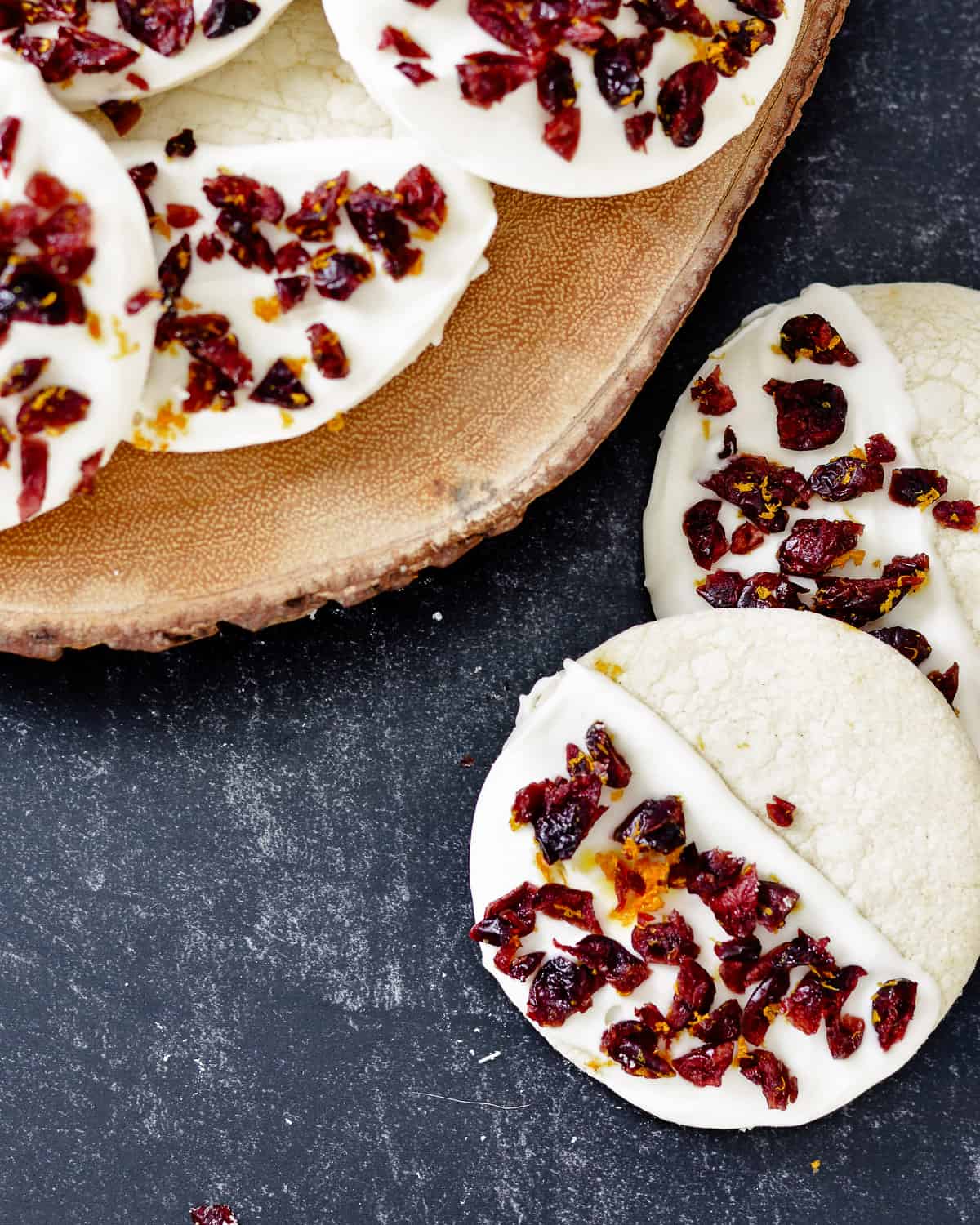 White chocolate dipped cookies with orange zest and cranberries on top, some on a wooden plate.