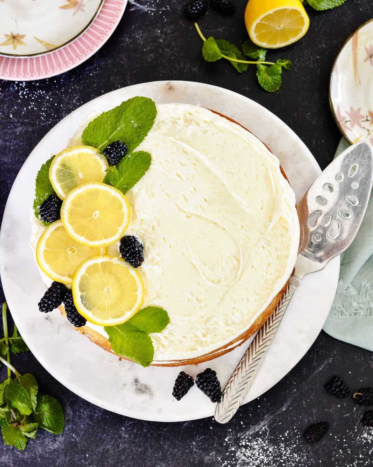 Lemons, blackberries and mint on top of frosting on a lemon cake. Cake server sitting next to cake.