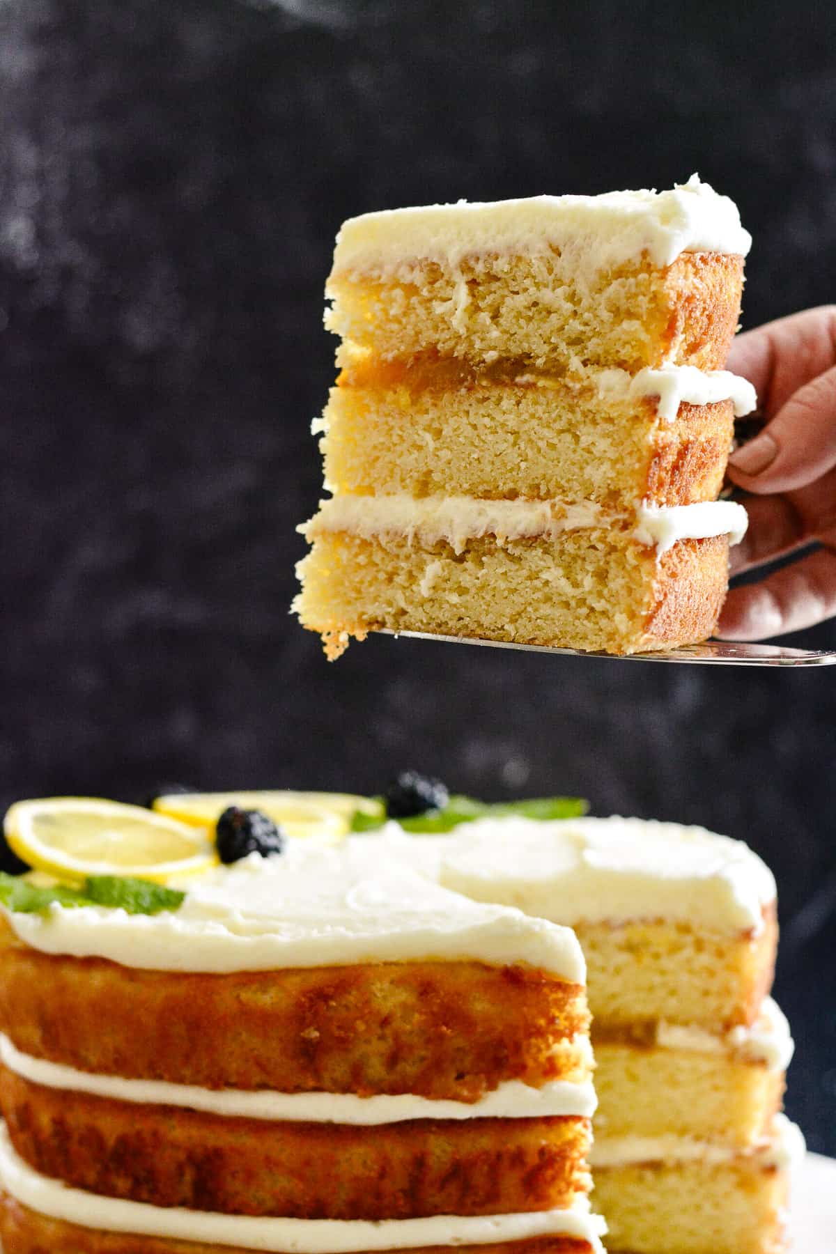 Hand holding a 3 layer slice of lemon cake on a cake server. Whole cake in the background on a cake plate.
