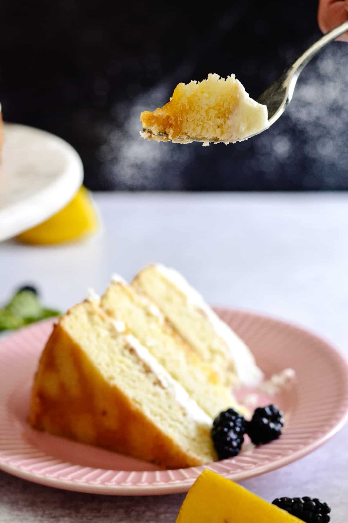 Fork with a bite of lemon cake with slice of cake in background.