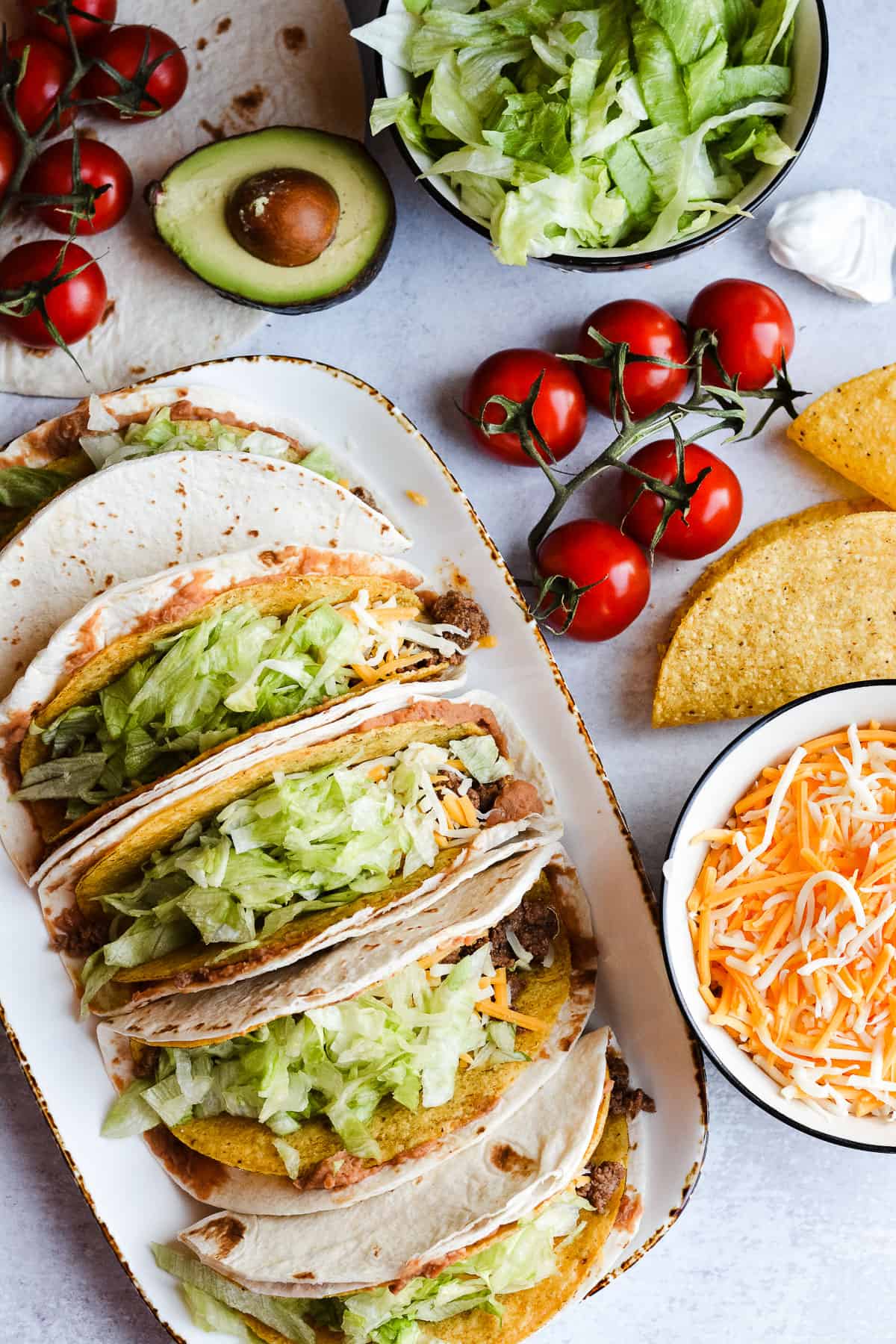 Plate of double decker tacos lined up with tomatoes, avocados and cheese around.