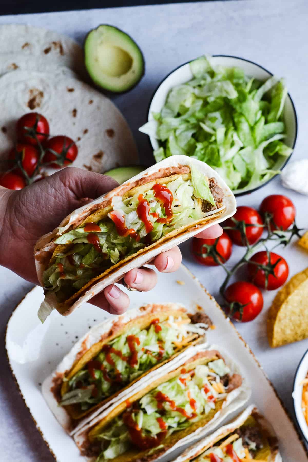 Hand holding taco with lettuce, avocado, tomatoes and taco shells in background.
