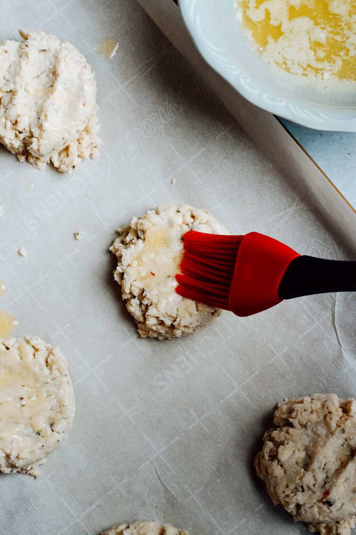 Scooped biscuit dough on baking sheet with pastry brush adding butter to tops.