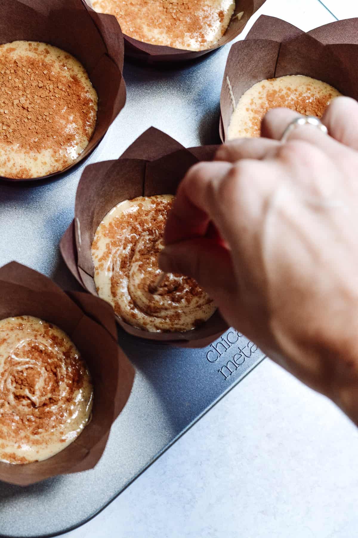 Hand using a toothpick to swirl in more cinnamon and raw sugar on top of cupcake batter.