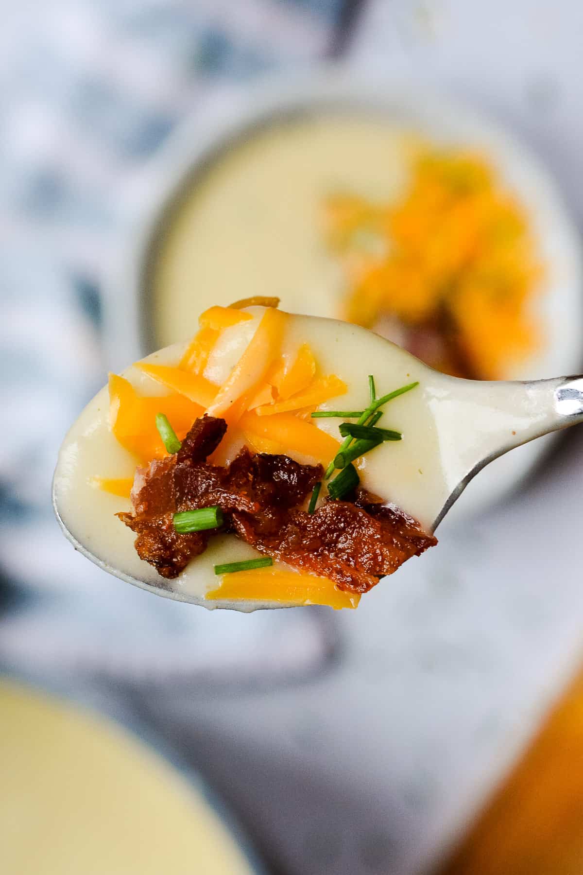 Potato soup on a spoon with cheddar cheese shreds, bacon bits and chopped chives. Bowl of soup and towel in background.