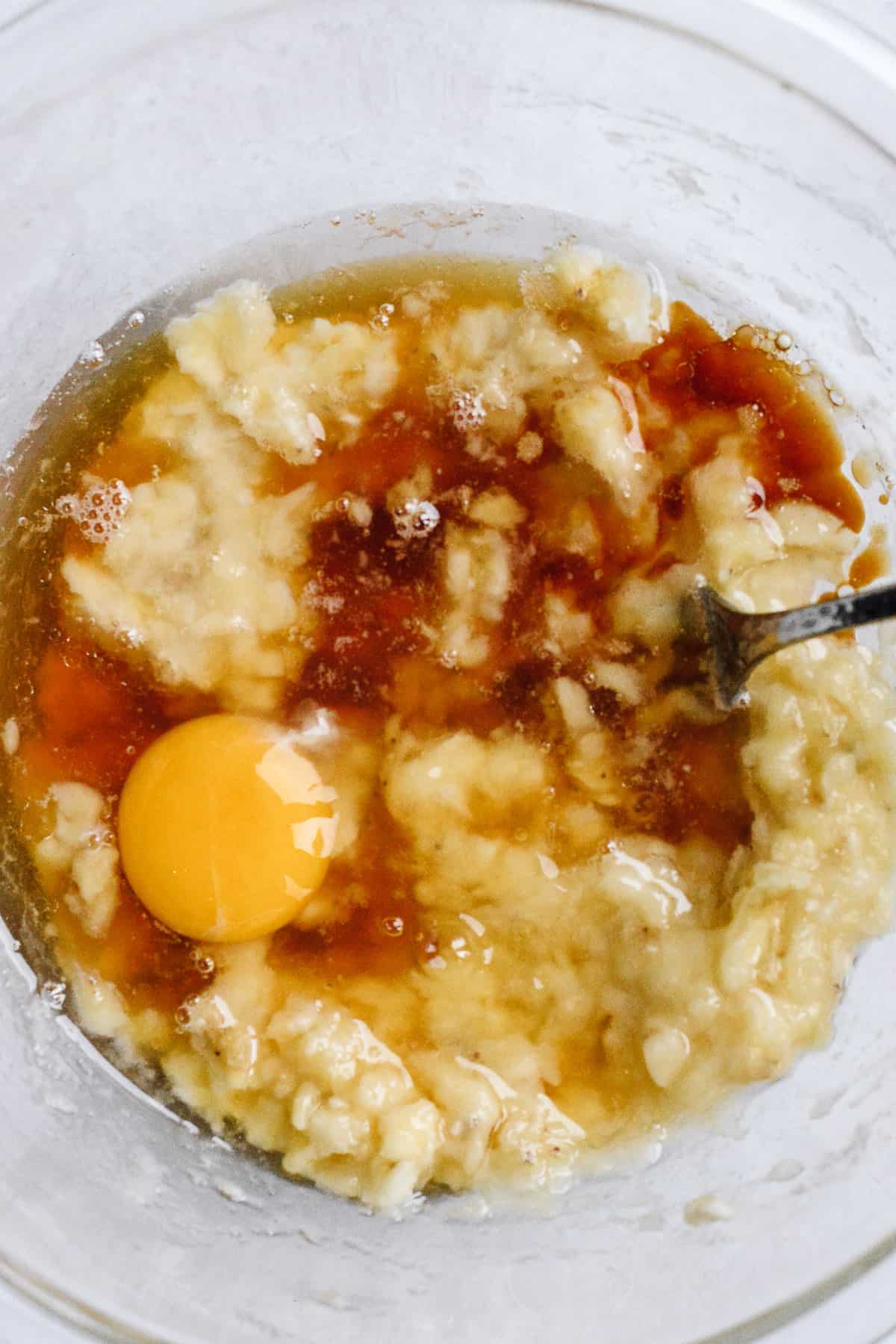 Egg, honey, vanilla and coconut oil added to mashed bananas in bowl with fork.