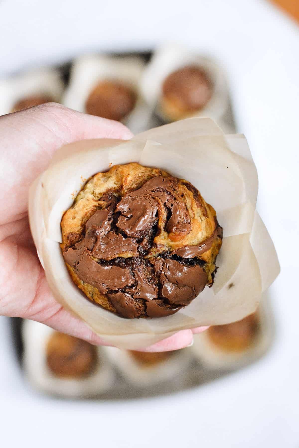 Hand holding up close of nutella banana muffin with tin full of muffins in background.