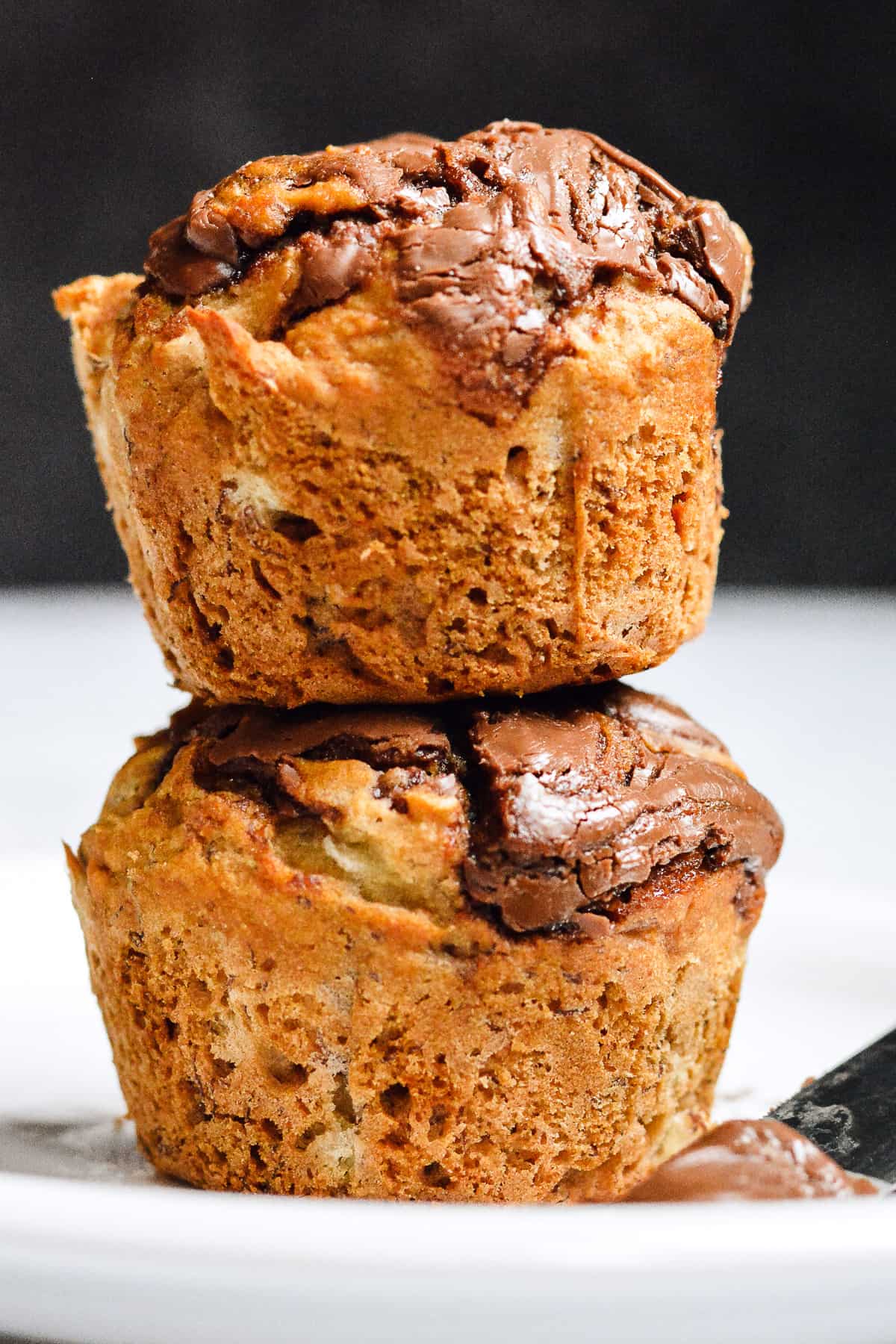 Two banana nutella muffins stacked on each other on top of white plate with nutella on knife in front of muffins.
