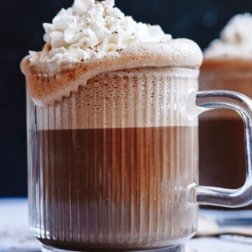 Hot chocolate in a glass mug with whip cream overflowing the edges.