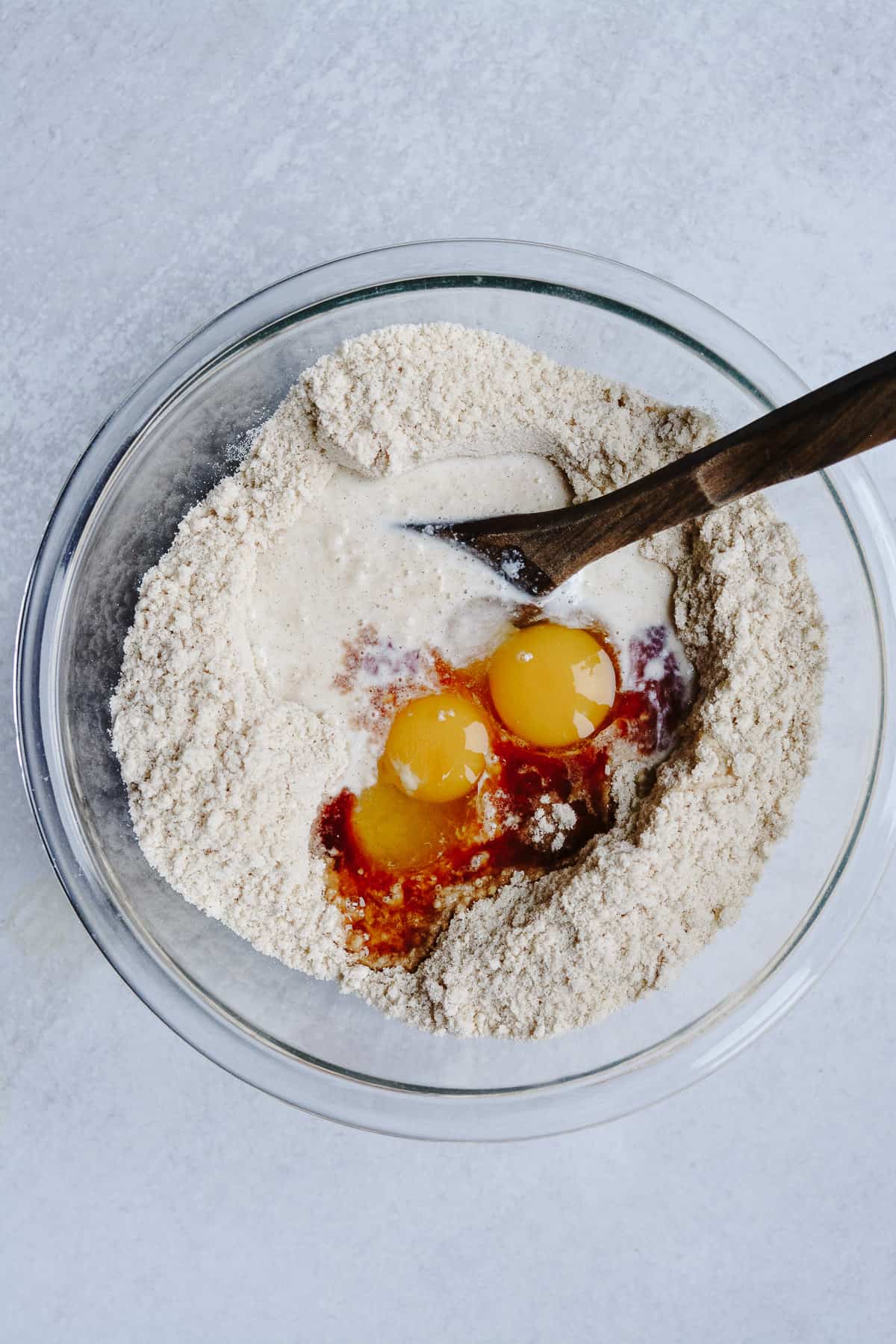 Dry ingredients with eggs, vanilla and milk in bowl with wooden spoon.