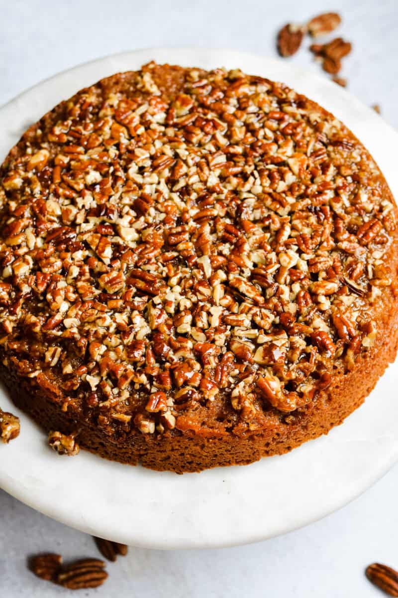 Whole pecan upside down cake on cake plate with raw pecans sprinkled around.