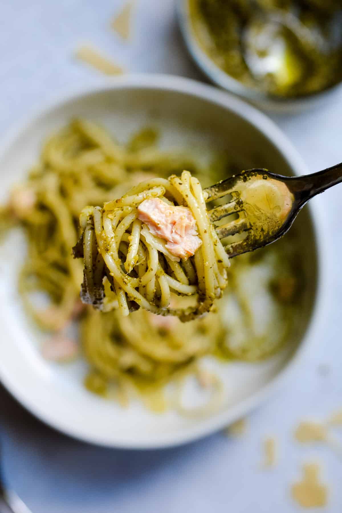 Close up of a fork with spaghetti, pesto and tuna, with bowl in background and pesto with spoon.