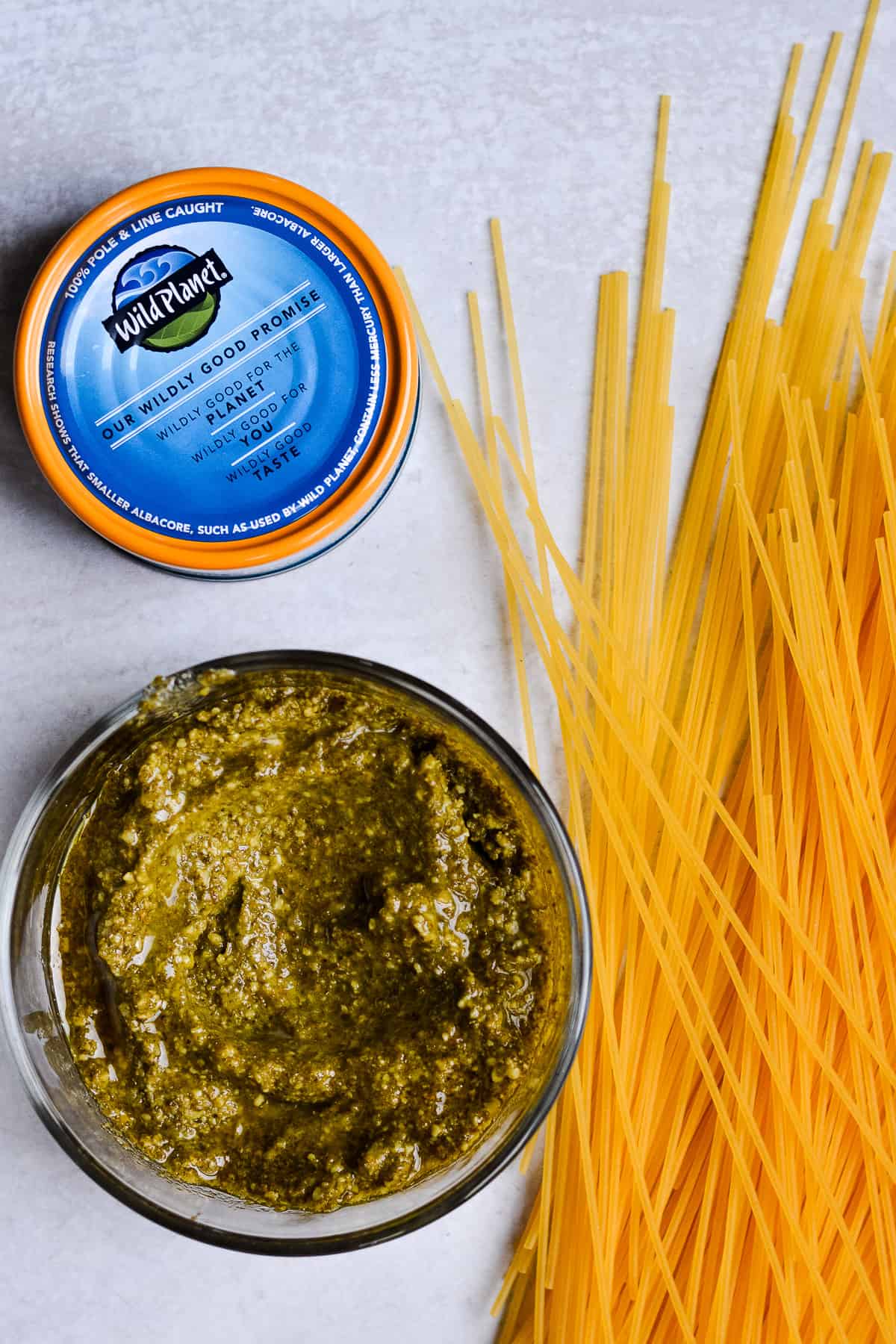 Spaghetti noodles, pesto in a bowl and can of Wild Planet tuna laying on a countertop.