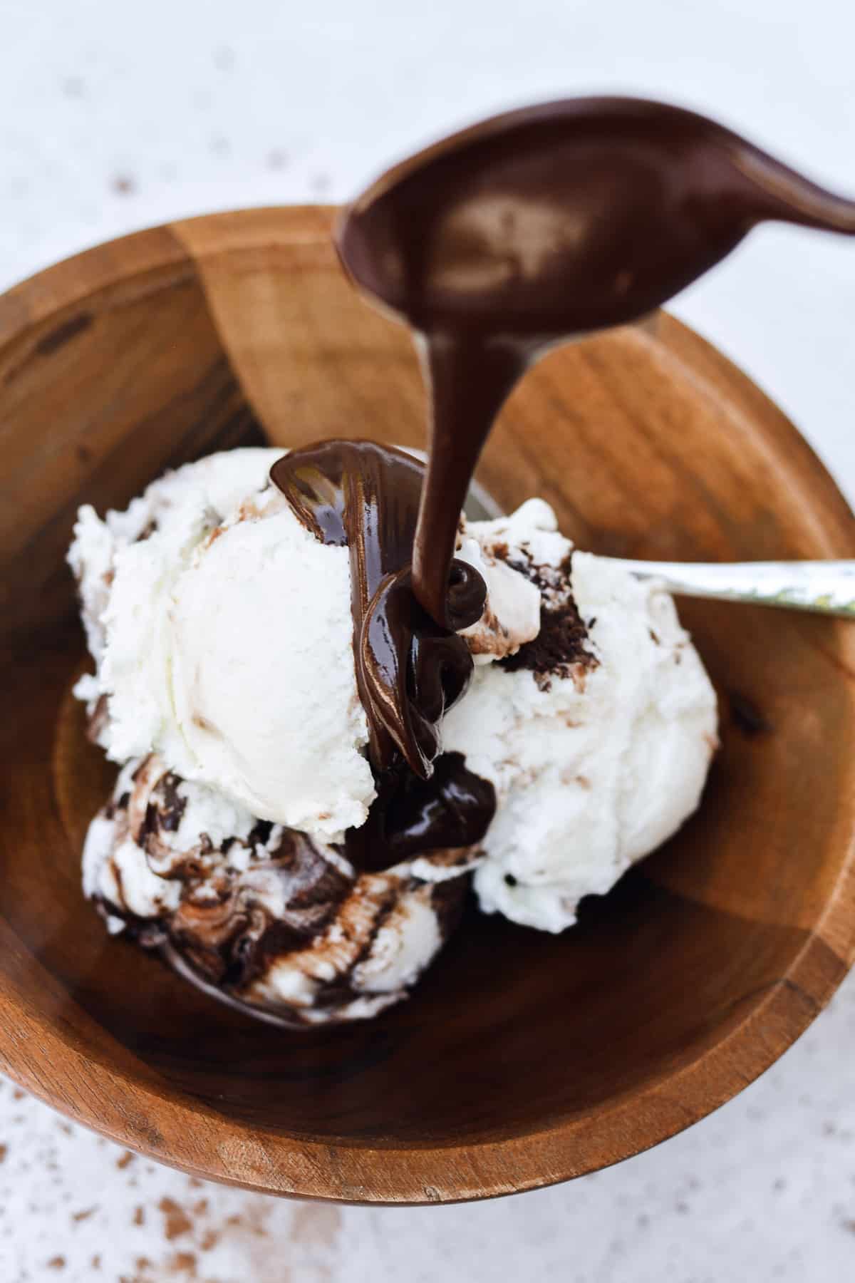 Spoon pouring sauce over vanilla ice cream in wooden bowl.