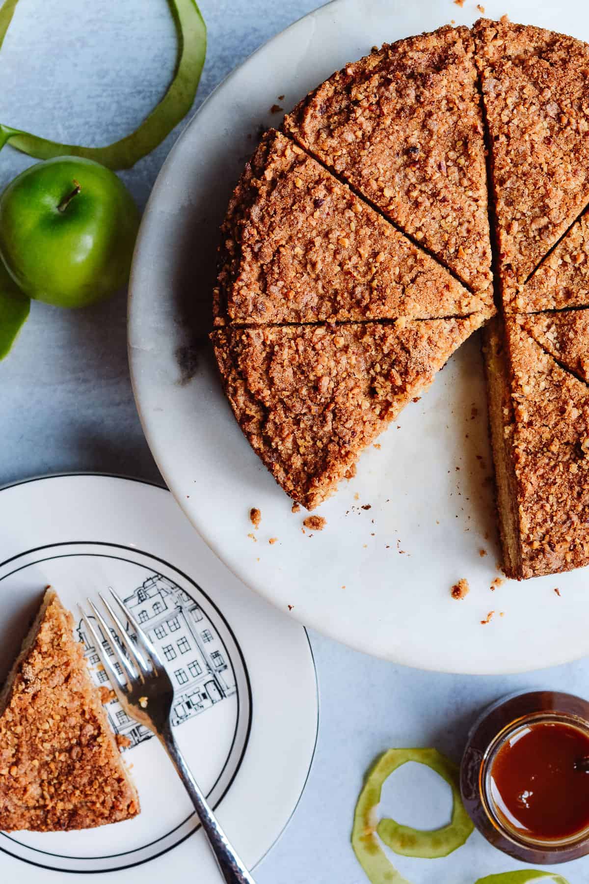 Close up of sliced apple cake with caramel sauce on the side and apple peel and apples around.